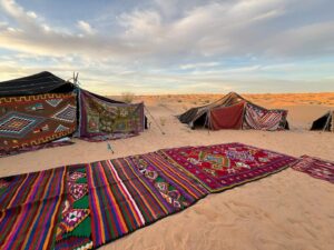 Tradizionale accampamento di tende beduine in tessuto scuro distese tra le dune di sabbia, con tappeti e sedute esterne nel deserto tunisino.