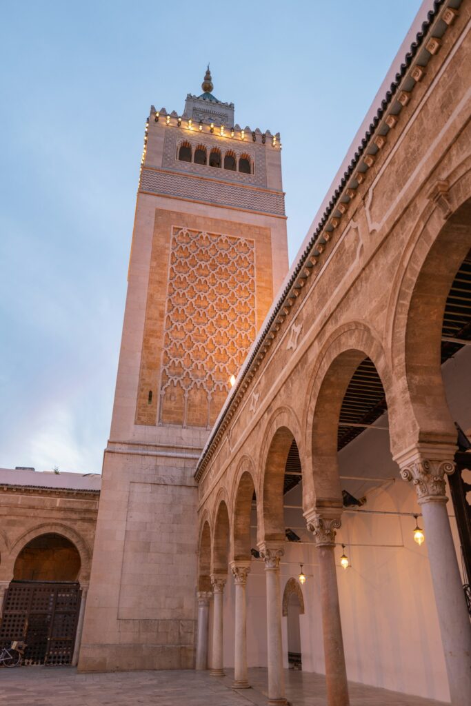 Cortile interno della Grande Moschea Al-Zaytuna a Tunisi, con portici ad arco, colonne antiche e il minareto decorato in stile almohade sullo sfondo.